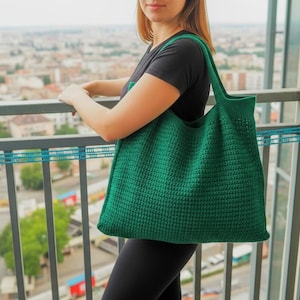 May include: A large, emerald green crochet tote bag with a textured stitch pattern. The bag has two long handles and is being held on a balcony overlooking a cityscape. The bag is made of a thick yarn.