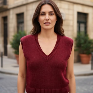 May include: A burgundy V-neck sweater vest. The sleeveless top is made of a knitted material. The woman is standing in front of a building with a street in the background. The vest has a ribbed hem and neckline.