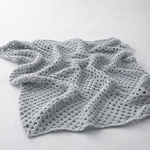 Könnte beinhalten: Eine hellgraue Häkeldecke mit einem strukturierten, offenen Webmuster. Die Decke ist gefaltet und drapiert, wodurch die komplizierte Sticharbeit sichtbar wird. Die weiche, neutrale Farbe und das offene Design deuten auf einen gemütlichen, handgefertigten Artikel hin.