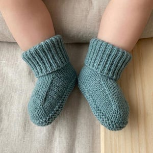 May include: Two teal knitted baby booties with ribbed cuffs and a textured knit design. The image shows the booties on a light-coloured surface. The booties are designed for babies, showcasing a focus on baby clothing and accessories.