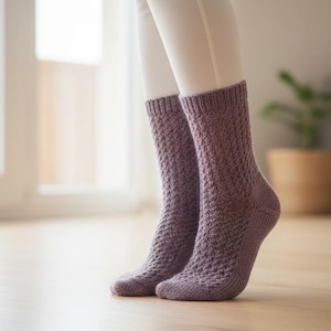 May include: A pair of lavender-colored knitted socks with a textured diamond pattern. The socks are worn on a person's legs, with the feet resting on a wooden floor. The background includes a window and a potted plant.