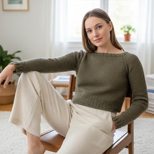 May include: An olive green sweater with long sleeves and a boat neck. The sweater has a textured knit pattern and is paired with cream-colored pants. The person is sitting on a wooden chair in a well-lit room.