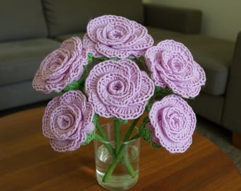 May include: A bouquet of handmade crochet flowers in a clear glass vase. The flowers are a soft lavender color with green stems and leaves. The crochet work is detailed, creating a realistic rose appearance. The arrangement sits on a wooden table.