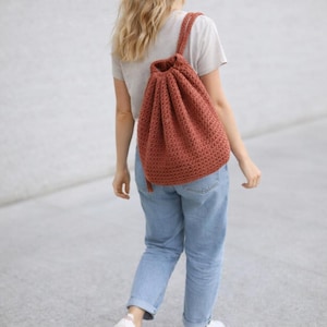 Könnte beinhalten: Ein rostfarbener, gehäkelter Rucksack mit Kordelzug und strukturiertem Stichmuster. Die Tasche hat einen Schultergurt und ein Quastendetail. Die Tasche wird über der Schulter getragen.