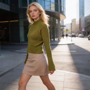 May include: A woman wearing a green ribbed knit sweater with long sleeves and a beige mini skirt. The woman is walking on a city street with buildings and cars in the background. The sweater has a high neckline.