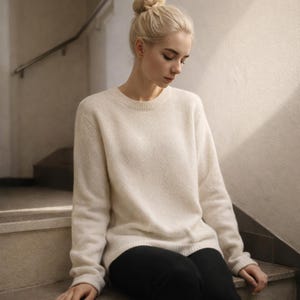 May include: A cream-colored, crew-neck sweater with long sleeves. The sweater has a soft, textured appearance. The person is wearing black pants. The background includes a staircase and a wall.