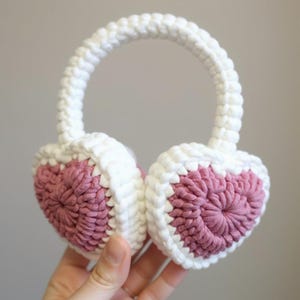 May include: Crochet earmuffs in a heart shape. The ear covers are a dusty rose colour, framed with white crochet. A white crochet band connects the ear covers. These earmuffs are designed for warmth and style.