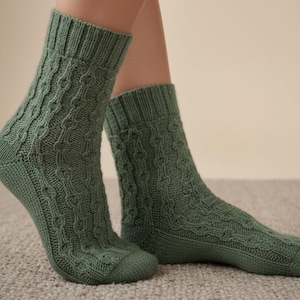 May include: A pair of green knitted socks with a cable knit pattern. The socks are made of a soft, textured material and have ribbed cuffs. The socks are displayed on a light-colored carpet.