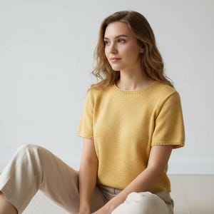 May include: A woman wearing a yellow short-sleeved knit top and beige pants. The top has a crew neck with a decorative stitch. The woman is sitting on a light-colored floor against a white background.