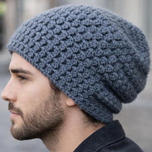 Puede incluir: Un gorro tejido a mano de color gris con un patrón tejido texturizado. El gorro está hecho de un material suave, posiblemente lana. El gorro es de corte relajado, adecuado para el frío. El sombrero es de color gris liso y apagado.