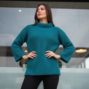 May include: Teal-colored ribbed knit sweater with a high neckline and flared sleeves. The sweater is paired with black pants. The model is standing in front of a glass wall.