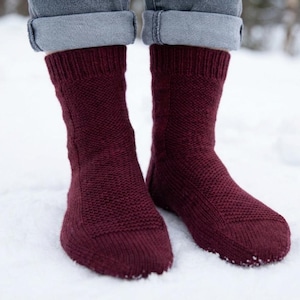 May include: Close-up of burgundy knitted socks worn with grey denim jeans. The socks, with a textured pattern, are visible from the ankles down. The scene is set in a snowy environment, with the socks standing in the snow.
