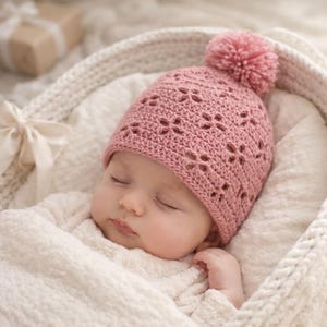 Puede incluir: Un bebé dormido con un gorro rosa tejido a ganchillo con un pompón. El gorro tiene un patrón floral y está en la cabeza del bebé. El bebé está envuelto en una manta blanca.