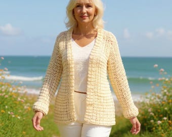 May include: A cream-coloured, open-front crochet cardigan with long sleeves. The cardigan is worn over a white top and paired with white trousers. The background features a beach scene with blue water and a clear sky.