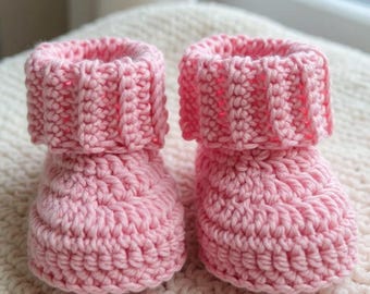 Op de afbeelding: Een paar roze gehaakte babyslofjes. De slofjes hebben een geribbelde manchet en een textuursteekpatroon. Ze worden gepresenteerd op een crèmekleurig, gestructureerd oppervlak, waarschijnlijk een deken of kleed. De zachte roze kleur suggereert een babyartikel.