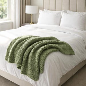 Peut inclure: Une couverture en tricot vert sauge drapée sur un lit blanc. La couverture présente un motif texturé et tissé. Le lit est composé de draps et d'oreillers blancs, avec une tête de lit de couleur neutre. Une lampe est posée sur une table de chevet à l'arrière-plan.