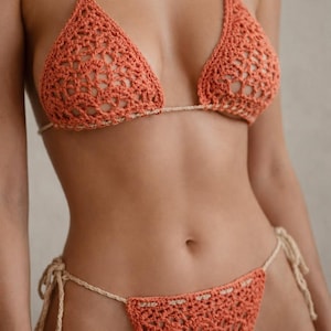 May include: A crocheted bikini set in a coral color. The top is a triangle style with a halter neck, and the bottoms are a string bikini. The edges of the bikini are a light beige color.