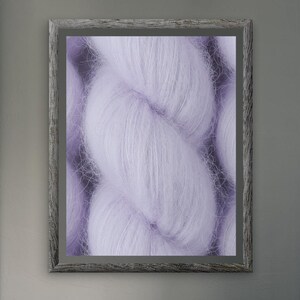 May include: A framed close-up of a braided skein of soft, fluffy, lavender yarn. The image is displayed on a gray wall, with a gray wooden frame. The room has gray curtains, a gray armchair, and a gray sofa with pillows.