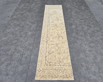 May include: A rectangular, beige runner rug with an intricate floral and geometric pattern. The rug features a border design and is placed on a gray surface. The rug's design includes floral motifs and decorative elements.