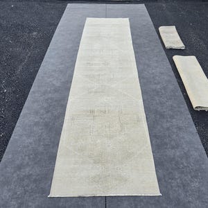 Peut inclure: Un long tapis de couloir étroit de couleur blanc cassé, orné d'un motif géométrique subtil et clair avec un dessin central en forme de croix. Deux sections plus petites et pliées du même tapis sont placées à droite du tapis principal.