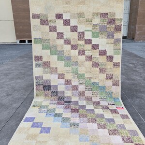 May include: A rectangular rug with a patchwork design. The rug features a repeating pattern of squares in various colors, including purple, green, and blue, set against a beige background. The rug is rolled out on a concrete surface.