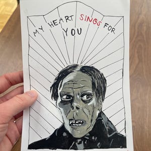 May include: Hand-painted artwork featuring a portrait of a character with gray skin and dark hair. The text "MY HEART SINGS FOR YOU" is written above the portrait in black and red ink. The artwork is on white paper.