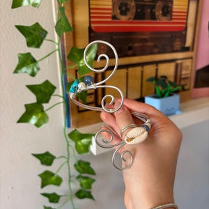 May include: A silver-toned wire cuff bracelet featuring two prominent spiral ends. One end is adorned with a white cowrie shell and a small blue bead, while the other end features a turquoise stone, a white pearl, and a small blue bead.