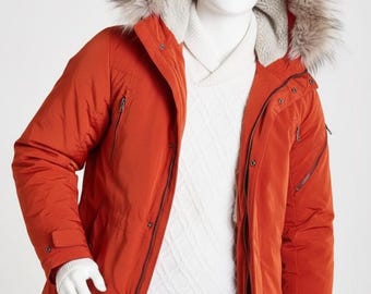 May include: A rust-colored winter parka with a faux fur-trimmed hood. The jacket features a zip closure, multiple pockets, and a white sweater underneath. The mannequin is also wearing gray pants and white gloves.