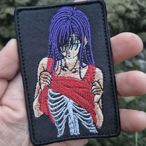 May include: Embroidered patch featuring an anime-style character with purple hair and blue eyes, wearing a red dress. The design reveals a skeletal torso. The patch is rectangular with a black border.