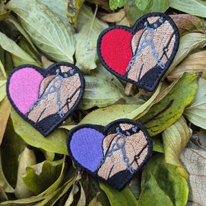 May include: Three heart-shaped embroidered patches with a black border. Each patch features a different color heart: pink, red, and purple. The other half of each heart has a tan design with blue chain details. Patches are on a bed of green and brown leaves.