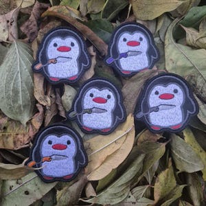 Könnte beinhalten: Gestickte Pinguin-Patches mit schwarzem Umriss, hellblauem Körper, roten Füßen und Schnabel sowie einem Messer. Die Patches sind auf einem Bett aus getrockneten Blättern verstreut. Der Pinguin hält ein Messer.