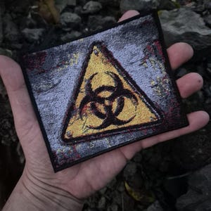 May include: Square patch with a yellow biohazard symbol inside a yellow triangle on a grey background. The patch has a distressed look with red and yellow accents. The edges are black.