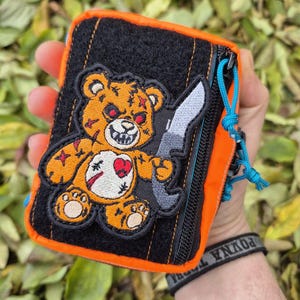 May include: An orange and black zippered pouch with a cartoon bear patch holding a knife. The bear has red eyes and a stitched heart. The pouch has a blue pull cord and a black zipper. The edges are trimmed in orange.