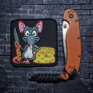 May include: An orange folding knife with a black lanyard and a skull bead next to a black patch. The patch features a cartoon rat holding a knife next to a block of cheese. The patch is square.