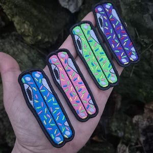 May include: Four colorful embroidered patches in blue, pink, green, and purple, each with a black border. The patches feature a stylized design with colorful sprinkles and a white and black element, resembling a butterfly knife.