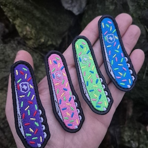 Könnte beinhalten: Vier farbenfrohe, gestickte Aufnäher in Form von Schweizer Taschenmessern. Jeder Aufnäher hat eine andere Farbe: lila, rosa, grün und blau, alle mit einem Streuseldesign. Die Aufnäher haben einen schwarzen Rand und eine weiße Messerklinge.