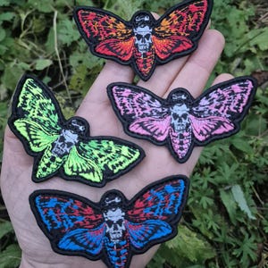 May include: Four embroidered moth patches in vibrant colors: red-orange, pink, blue-red, and green. Each patch features a skull design on the moth's body. The patches have a black border and are displayed against a backdrop of green foliage.