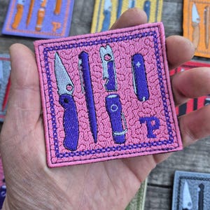 May include: Embroidered pink patch featuring a variety of purple tools, including a knife, pen, and multi-tool. The patch has a purple border and a letter "P" in the lower right corner. Other patches with similar designs are visible in the background.