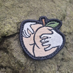 May include: Embroidered patch featuring a peach with hands. The peach is light brown with a green leaf and orange stem. White hands are on the peach. The patch has a black border.