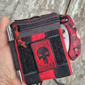 May include: A black and red tactical pouch with a skull logo and the text "POVNA TORBA". A silver pen is in a side pocket. A red and black knife is visible.