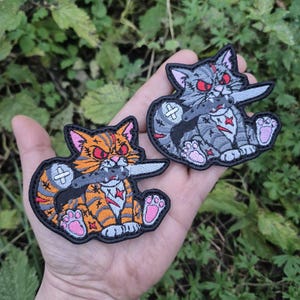 May include: Two embroidered cat patches. One is orange and black, the other grey and white. Both have red eyes and hold knives. The patches have pink paw pads and are set against a green background. The patches are suitable for clothing.