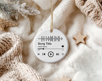 May include: A white, round ornament with a gold string for hanging. The ornament features a music player interface design with the text "Song Title" and "Artist Name". A heart icon and wooden star decorations are also visible.