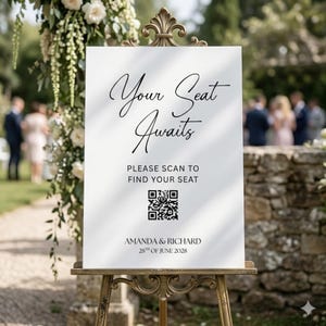 May include: A white wedding sign on a decorative gold easel. The sign reads "Your Seat Awaits" in elegant script, with the text "Please scan to find your seat" below a QR code. The sign also includes the names "AMANDA & RICHARD" and the date "28th of June 2028".