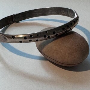 May include: A silver-toned bangle bracelet with a series of small, evenly spaced holes. The bracelet rests on a smooth, rounded, brown stone. The bracelet has a simple, elegant design.