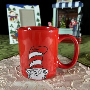 May include: A red ceramic mug featuring a cartoon cat wearing a tall, striped hat. The mug has a curved handle and a glossy finish. The design is a popular children's book character. Other decorative items are in the background.