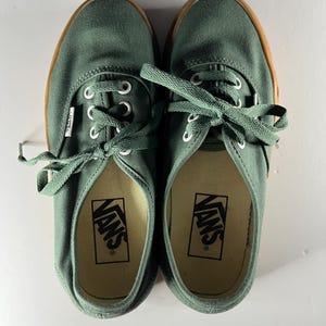 以下が含まれることがあります： 白い靴紐と茶色のラバーソールが付いた、グリーンのキャンバスVansスニーカーのペア。 Vansのロゴがインソールに見えます。 上から見た図。