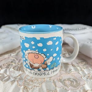 May include: A white ceramic mug with a blue interior and handle. The mug features a cartoon illustration of a character in a winter scene with the text "GIVE US GRATEFUL HEARTS". The mug has a white rim with blue polka dots.