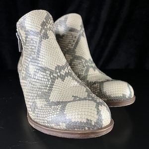 May include: Ankle boots with a snakeskin pattern in grey and cream. The boots have a side zip and a brown sole. The boots are made of a textured material. The boots are a fashionable footwear item.