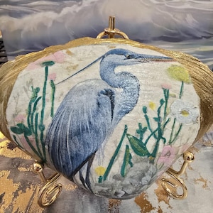 May include: A decorative seashell featuring a blue heron illustration. The shell has a gold-painted edge and is supported by a gold-colored metal stand. The heron is depicted amidst colorful flowers and greenery.
