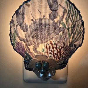 May include: A decorative night light featuring a seashell design with a crab illustration. The shell is adorned with blue, green, and purple accents. The base is white, and the light is illuminated. The night light is decorated with a flower and blue pearls.